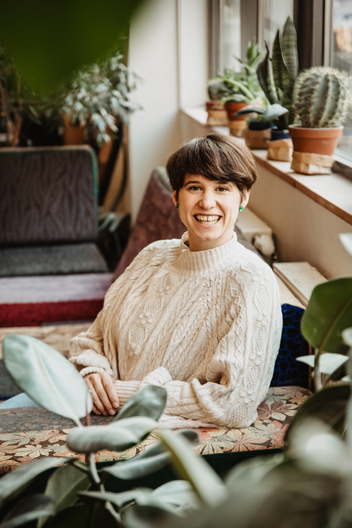 Clara Fohrbeck unterwegs für therapeutisches Klettern und Systemisches Coaching sitzt in einer Boulderhalle in Berlin in weißem Pulli auf Sofa zwischen Pflanzen und lächelt in die Kamera.